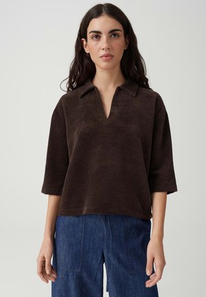 Vrouw met lang donker haar die een bruine geribde blouse met kraag en driekwart mouwen draagt en een blauwe spijkerbroek tegen een effen achtergrond.