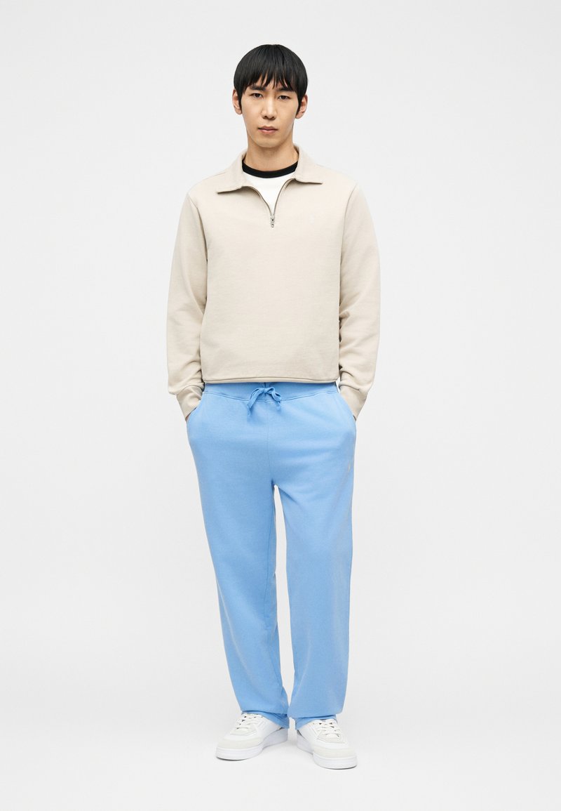 Jeune homme debout les mains dans les poches d’un pantalon de survêtement bleu clair, portant un sweat-shirt beige à fermeture éclair quart de zip et des baskets blanches sur fond blanc.