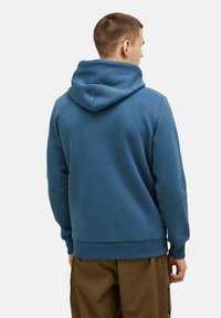 Felpa blu realizzata in morbido tessuto, con cappuccio regolabile e polsini a coste, abbinata a pantaloni marroni, dimostrando un design semplice e funzionale.