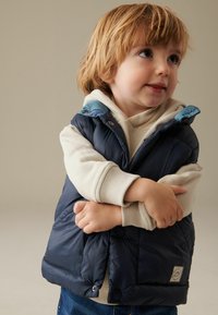 Gilet imbottito blu navy con colletto azzurro e felpa crema sotto; presenta chiusure a bottone e tasche laterali.