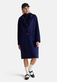 Cappotto in lana blu navy con ampi revers, due tasche e bottoni grandi. Indossato sopra un dolcevita nero, abbinato a calzini bianchi e scarpe nere.