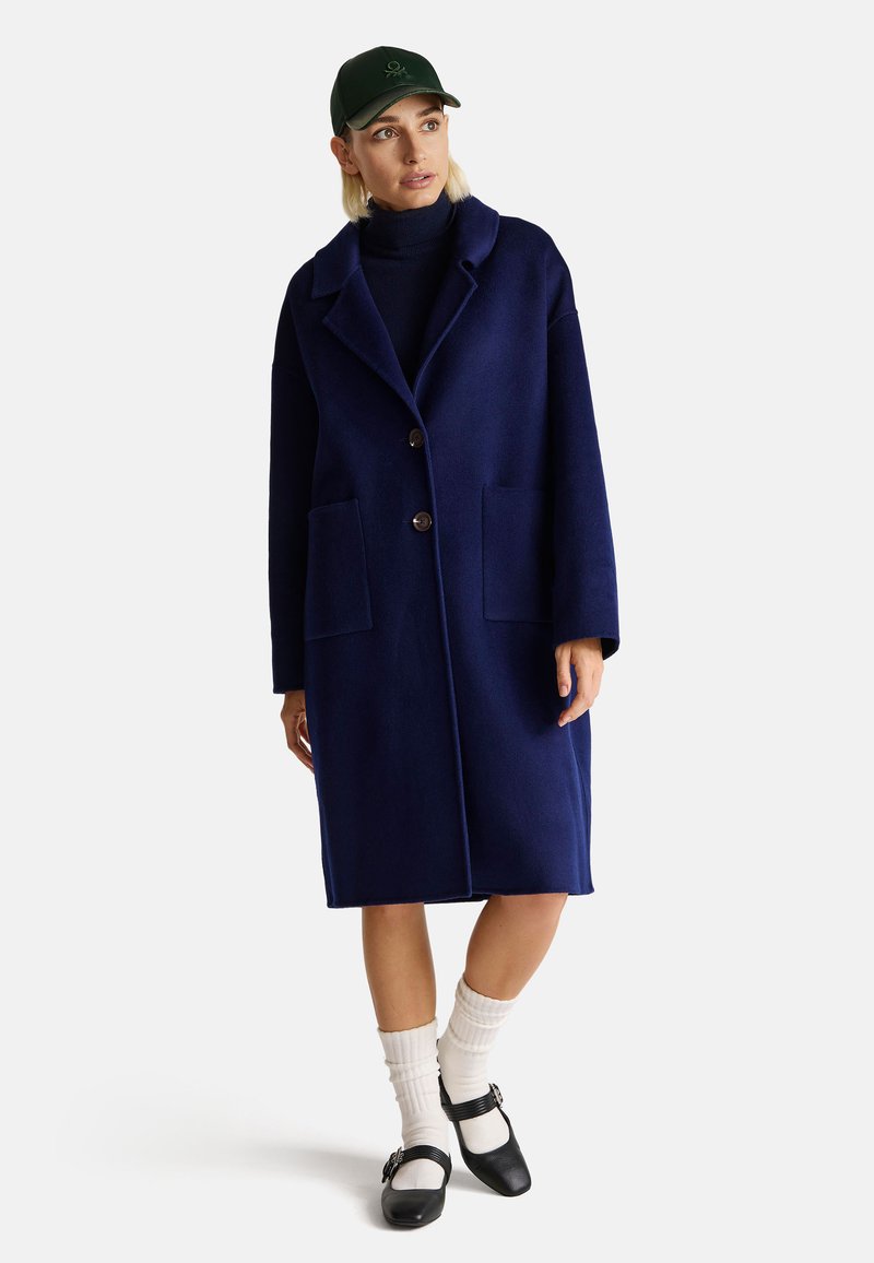 Cappotto in lana blu navy con ampi revers, due tasche e bottoni grandi. Indossato sopra un dolcevita nero, abbinato a calzini bianchi e scarpe nere.