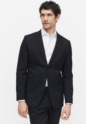 Man in een zwart pak met een wit overhemd, staand tegen een effen witte achtergrond, kijkend naar de zijkant.