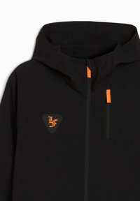 Sort hættejakke lavet af glat stof, med orange lynlåstræk og et trekantet logo mærke med orange skrift.