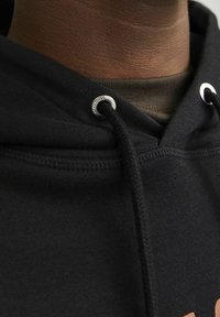 Sudadera negra con capucha, que cuenta con un cordón y ojales de metal, de textura suave, superpuesta sobre una camisa marrón con un patrón acanalado.