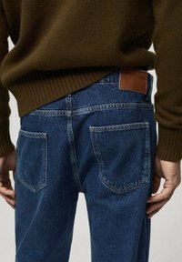 Jeans de mezclilla azul con corte recto, que cuentan con dos bolsillos traseros y un parche de cuero marrón en la cinturilla. Parte de una blusa de punto acanalado es visible.