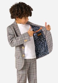 Completo grigio a fantasia a scacchi con bottoni chiari. L'interno è caratterizzato da un tessuto scuro con design geometrici colorati. Camicia bianca sotto.