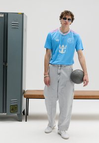 Camisola desportiva azul claro com riscas e logótipo rosa, combinada com calças largas de ganga cinzentas. Modelo a segurar uma bola preta, em pé junto a um cacifo.