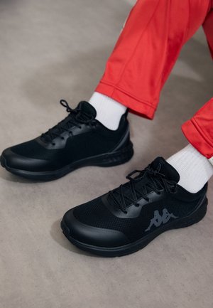 Pies con zapatillas deportivas negras, calcetines blancos y pantalones rojos, de pie sobre un suelo gris y liso.