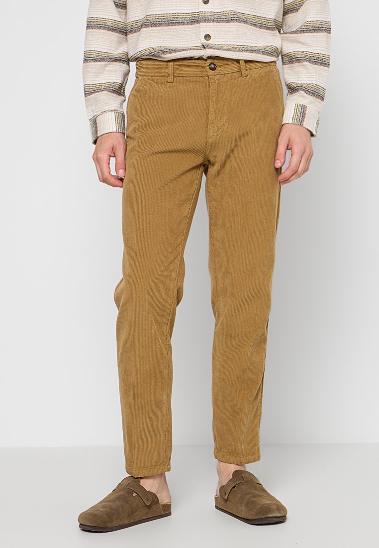 Pantaloni in velluto corduroy marroni con vestibilità dritta, caratterizzati da una superficie testurizzata, tasche frontali e chiusura a bottone. Indossati con scarpe marroni senza lacci.