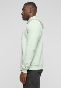 Felpa verde menta con tasca a marsupio, polsini a coste e cappuccio con cordino, realizzata in tessuto morbido. Il modello indossa jeans neri.