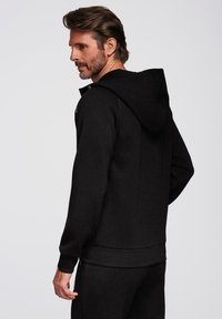 Sudadera negra hecha de tela suave, con una gran capucha, un bolsillo tipo canguro y puños y dobladillo acanalados. Los pantalones de chándal combinan con la sudadera.