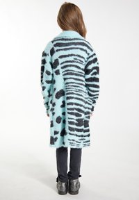 Dlhý, nadrozmerný sveter v svetlom modrom odtieni s kombináciou čiernych pruhov a bodkovaných vzorov. Mäkšia, nadýchaná textúra; nosený cez čierne nohavice.