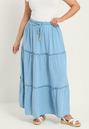 Kvinde iført en lyseblå flerlags maxi nederdel med snøre i taljen, kombineret med hvide sandaler og en hvid top med korte ærmer.