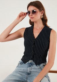 Námornícka bezrukávová vesta s bielymi pruhmi, s hlbokým výstrihom do V a šiestimi čiernymi gombíkmi. Skombinované s svetlomodrými vysokými džínsami.