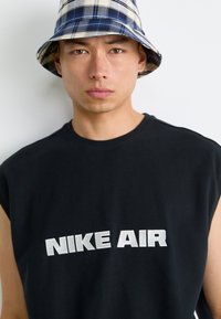 Mlad moški nosi modro-bež karo klobuk in črno majico brez rokavov s napisom "NIKE AIR" v beli barvi.