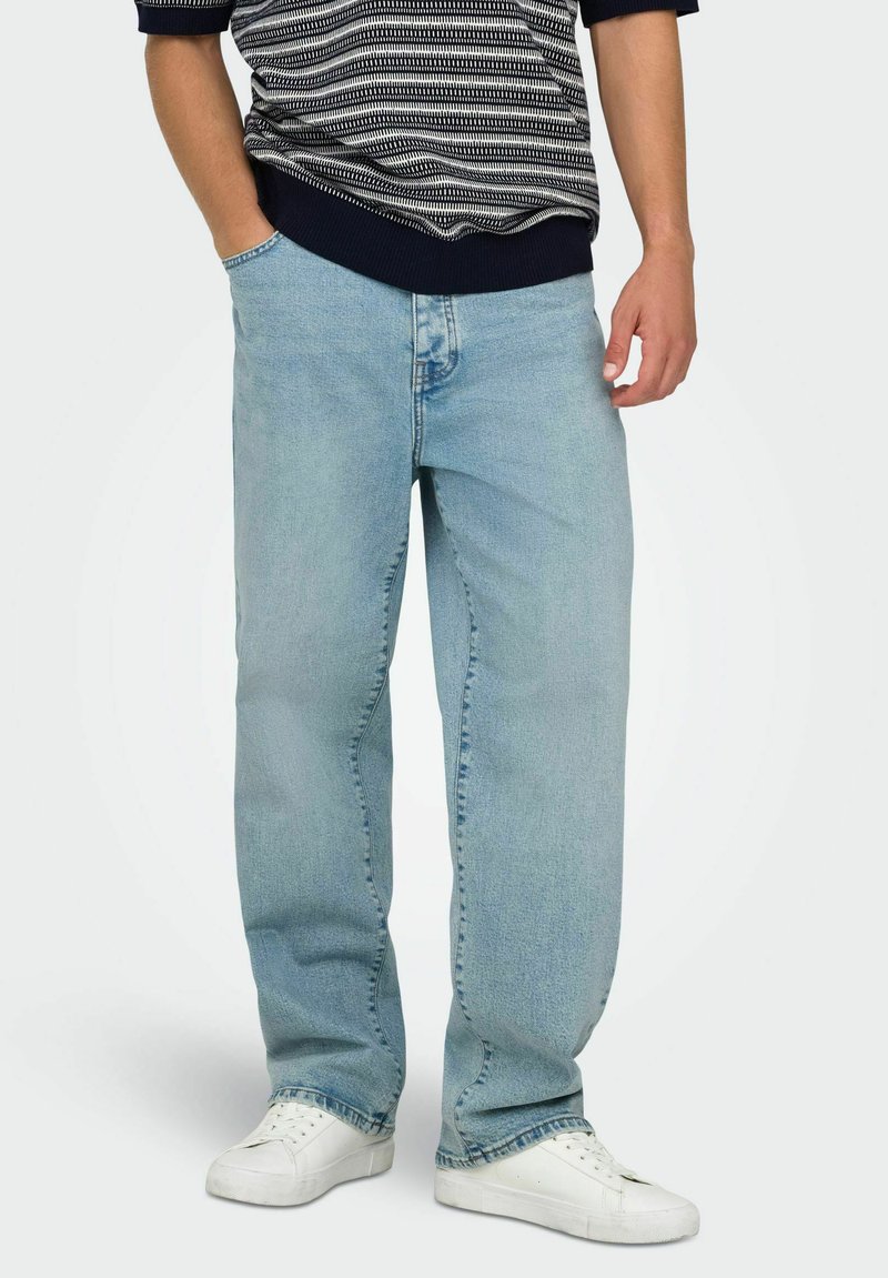 Personne portant un jean bleu clair droit, des baskets blanches et une chemise à manches courtes noire et blanche à motif, les mains détendues.