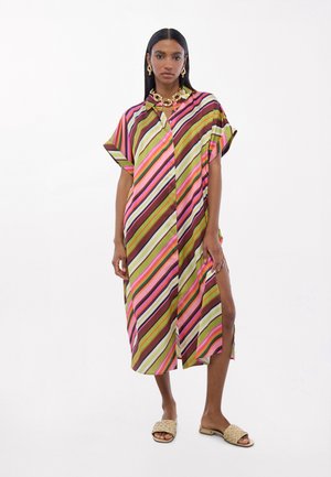 Mujer que lleva un vestido suelto a rayas diagonales de colores, con mangas cortas, abertura lateral, collar de cadena dorada y pendientes, y sandalias tejidas tipo slide.