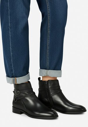 Sorte læderankelstøvler med sidelås og spænder båret med opsmøgede mørkeblå jeans på hvid baggrund.