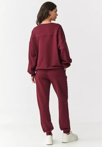 Bordowy zestaw bluzy i spodni dresowych wykonany z miękkiego materiału. Luźny krój, ściągacze przy rękawach i w pasie. Białe sneakersy uzupełniają stylizację.