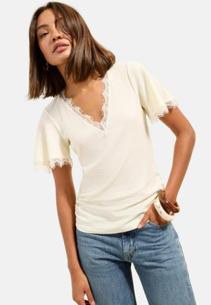 Frau trägt cremefarbenes, kurzärmeliges Spitzenoberteil mit V-Ausschnitt, blaue Jeans und klobige Armbänder am linken Handgelenk vor schlichtem Hintergrund.