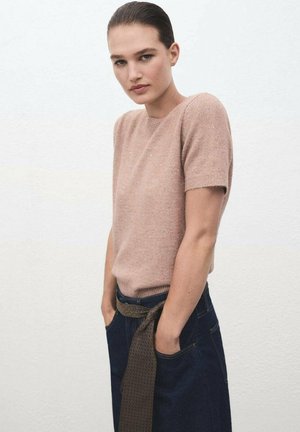 Jonge vrouw met slicked-back haar, een lichtbruine trui met korte mouwen en donkere broeken met een gestructureerde riem, handen in de zakken.