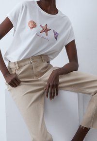 Vit bomullströja med skaldjurs- och sjöstjärnemönster i korall, orange, lila och texten "Seashell Beach Club." I kombination med beige högmidjade jeans.