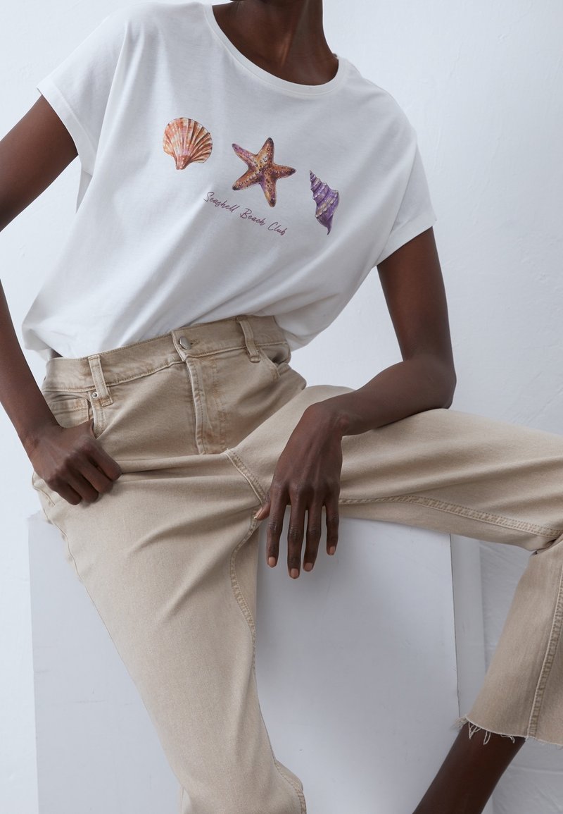 Vit bomullströja med skaldjurs- och sjöstjärnemönster i korall, orange, lila och texten "Seashell Beach Club." I kombination med beige högmidjade jeans.