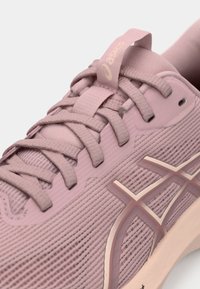 Primer plano de una zapatilla de malla rosa de ASICS con cordones, etiqueta de tela en la lengüeta y logo en el lateral, mostrando detalles de tejido texturizado y costuras.