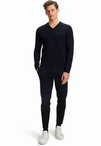 Granatowy sweter z długim rękawem i dekoltem w serek, zestawiony z pasującymi joggerami. Tkanina wydaje się dziana o gładkiej teksturze. Stylizację dopełniają białe trampki.