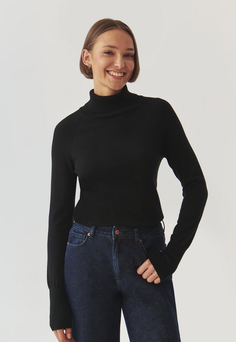 Maglione a collo alto nero con design aderente, colletto a coste e maniche lunghe, abbinato a jeans in denim scuri. Tessuto liscio, dettagli minimi.