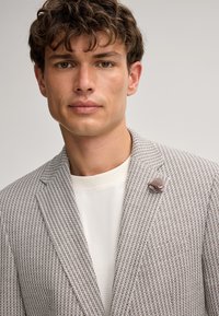 Hellgrauer Blazer mit strukturiertem Muster, Ein-Knopf-Verschluss, Reverskragen und einer Stoffblume als Anstecker. Darunter ein weißes T-Shirt mit Rundhalsausschnitt.