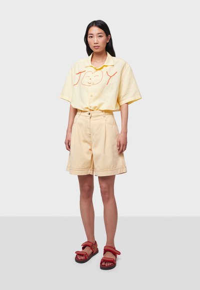Chemise jaune à manches courtes avec broderie rouge "JOY", associée à un short beige et des sandales rouges. Design simple et décontracté avec une coupe ample.