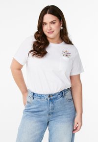 Bílý bavlněný tričko s krátkými rukávy a kapsou na hrudi, který má barevné květinové vyšívání; spojené se světle modrými džíny s vysokým pasem.