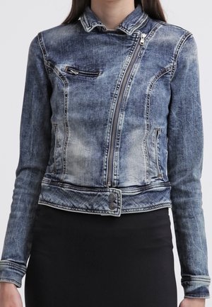 Chaqueta vaquera azul descolorida con cremallera asimétrica frontal, bolsillos en el pecho y laterales con cremallera, cinturilla con botones, llevada sobre una falda negra.