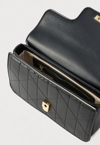 Musta tikattu nahkalaukku kullanvärisellä lukolla, avoinna ja esillä beige vuori sekä sisällä vetoketjullinen tasku.