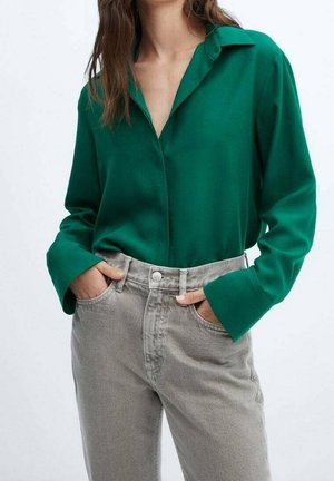 Femme portant un chemisier vert ample à manches longues, rentré dans un jean taille haute gris clair, les mains dans les poches.