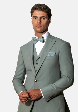 Homme portant un costume trois pièces vert clair avec un nœud papillon à motif et une pochette, ajustant les boutons de sa veste.