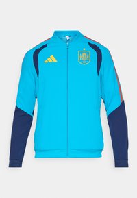 Türkizkék sportdzseki tengerészkék ujjakal, sárga cipzárral és Adidas logóval. A spanyol nemzeti címer a mellkason látható.