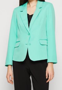 En kvinna som bär en figursydd turkos kavaj med två knappar och fickor med lock över en svart topp och svarta byxor, mot en enfärgad bakgrund.