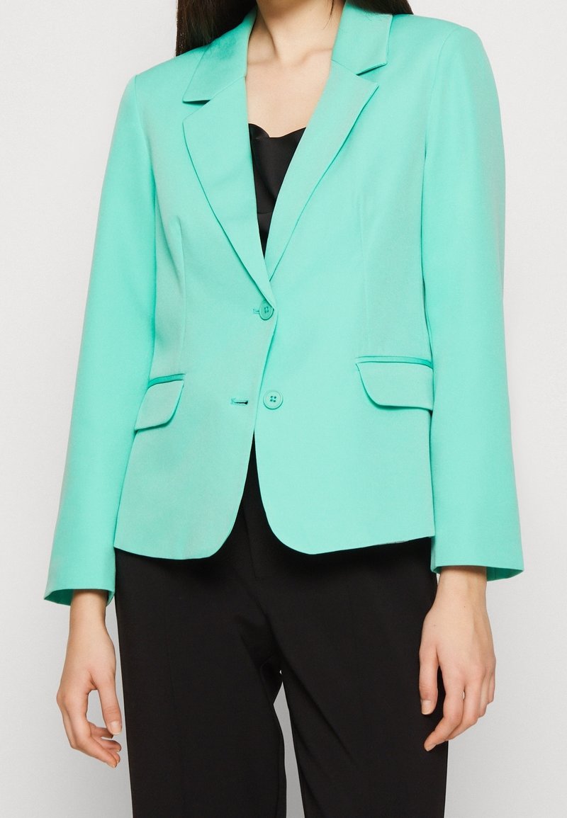 En kvinna som bär en figursydd turkos kavaj med två knappar och fickor med lock över en svart topp och svarta byxor, mot en enfärgad bakgrund.