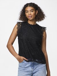 Femeie cu păr creț poartă un top negru fără mâneci din dantelă și blugi bleu deschis, stând cu o mână în buzunar, pe un fundal alb.