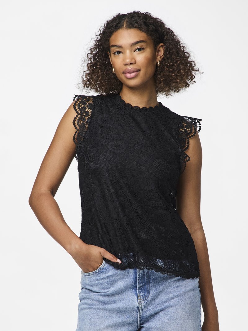 Femeie cu păr creț poartă un top negru fără mâneci din dantelă și blugi bleu deschis, stând cu o mână în buzunar, pe un fundal alb.