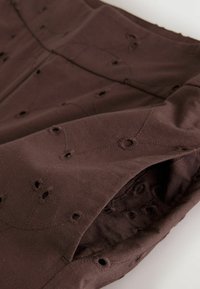 Närbild av brun tyg med broderade öljetter i blommiga och geometriska mönster på en vikt plaggärm.
