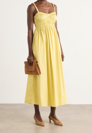 Kvinde iført en gul midi-kjole uden ærmer, med en brun clutch i hånden og fletvævede brune hæle, stående mod en ensfarvet lys baggrund.