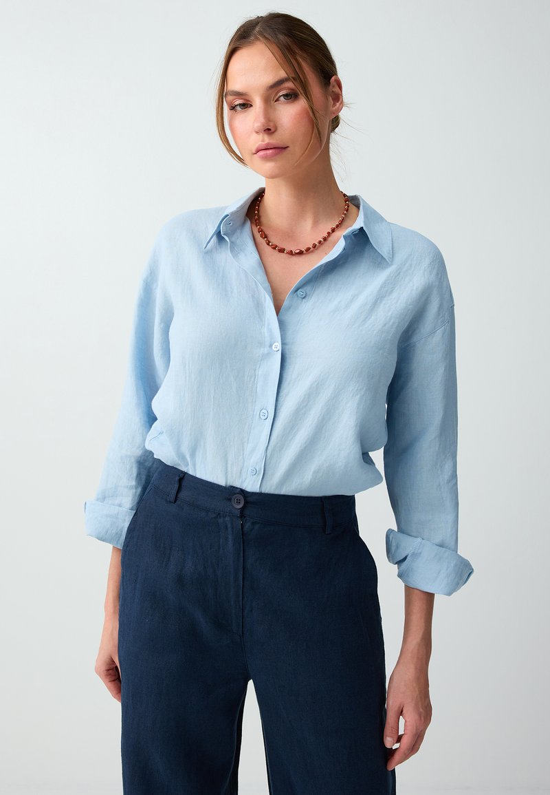 Femme portant une chemise bleu clair boutonnée aux manches retroussées, un pantalon bleu foncé et un collier de perles rouge, debout devant un fond uni.
