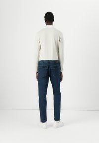 Camisa de manga larga de color claro, jeans azul oscuro y zapatillas blancas. Tejido suave y corte clásico, con los bolsillos traseros visibles.