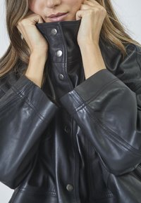 Giacca di pelle nera con colletto alto, bottoni a scatto e maniche larghe. Texture liscia e design minimalista con dettagli di cucitura visibili.