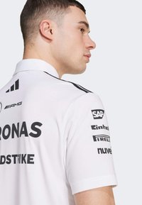 White polo shirt with short sleeves, featuring black logo prints on the back and shoulders, and a two-button collar. Smooth fabric texture.