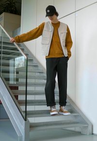 Outfit casual compus dintr-o vestă bej deschis, un pulover muștar, pantaloni închisi și adidași maronii, pozează pe o scară cu balustradă din sticlă.